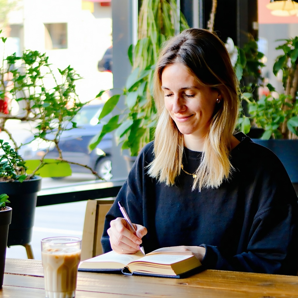 Woman journaling in coffee shop with plants and a window nearby
